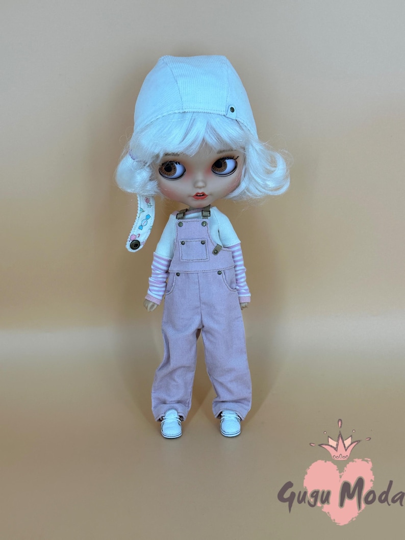 May include: A doll wearing a white corduroy hat, white wig, and pink overalls. The doll has large brown eyes, a white shirt with pink striped sleeves, and white sneakers. The doll is standing against a light brown background. The doll is wearing a white corduroy hat.