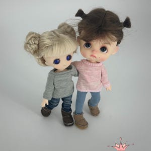 May include: Two handmade dolls with detailed features. One doll has blonde hair in a bun, wearing a gray sweater, jeans, and brown boots. The other doll has brown hair in pigtails, wearing a pink sweater, blue jeans, and brown boots. The dolls are standing together.