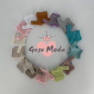 May include: A collection of 12 miniature knitted sweaters in various colors, arranged in a circle around a pink heart with the text "Gugu Moda" and a crown.