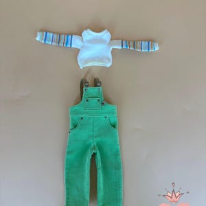 May include: A doll-sized outfit featuring a white long-sleeved shirt with striped sleeves and green corduroy overalls. The overalls have pockets and adjustable straps. The outfit is designed for a doll.