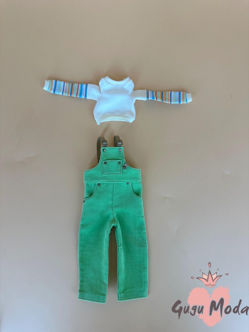 May include: A doll-sized outfit featuring a white long-sleeved shirt with striped sleeves and green corduroy overalls. The overalls have pockets and adjustable straps. The outfit is designed for a doll.