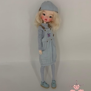 Può includere: Una bambola con capelli biondi indossa un berretto azzurro, una maglia a maniche lunghe a righe e un vestito a salopette in stile denim azzurro con un ricamo a farfalla viola sul petto. La bambola indossa anche leggings a righe e scarpe azzurre.