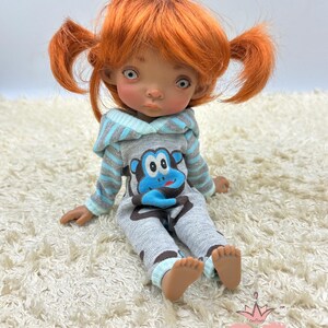 May include: A small doll with orange hair in pigtails wearing a gray and blue hooded jumpsuit with a cartoon monkey design. The doll is sitting on a white fluffy rug.