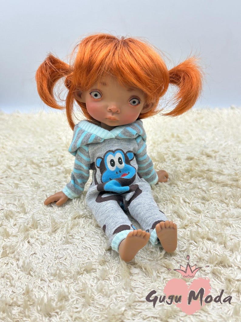 May include: A small doll with orange hair in pigtails wearing a gray and blue hooded jumpsuit with a cartoon monkey design. The doll is sitting on a white fluffy rug.