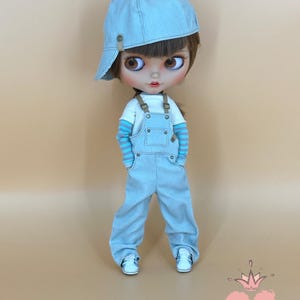 May include: A doll wearing a light blue denim cap, overalls, and white sneakers. The doll has brown hair and large brown eyes. The doll's shirt has blue and white striped sleeves. The doll is standing in front of a light brown background.