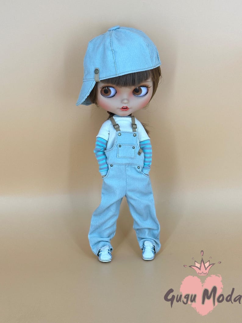 May include: A doll wearing a light blue denim cap, overalls, and white sneakers. The doll has brown hair and large brown eyes. The doll's shirt has blue and white striped sleeves. The doll is standing in front of a light brown background.