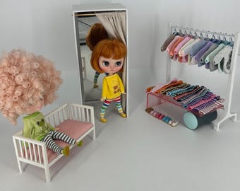May include: Two dolls with pink and blonde hair wearing striped pajamas and a yellow shirt with a bear graphic. The dolls are sitting on a white bed and a white bench. There is a white closet with a mirror and a white clothing rack with colorful clothes hanging on it. The clothing rack is on a white table with a clear glass top. There are colorful bracelets and shoes on the table. The background is white.