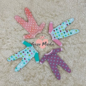 May include: Four baby doll onesies in different colors and patterns. Each onesie has a heart print with different colored hearts. The onesies are pink, light blue, turquoise, and purple.