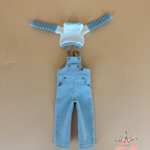 May include: A light blue corduroy overall set with a white and blue striped long-sleeve shirt. The overall has pockets and adjustable straps. The set is displayed on a tan background. The text "Gugu Moda" is in the bottom right corner.