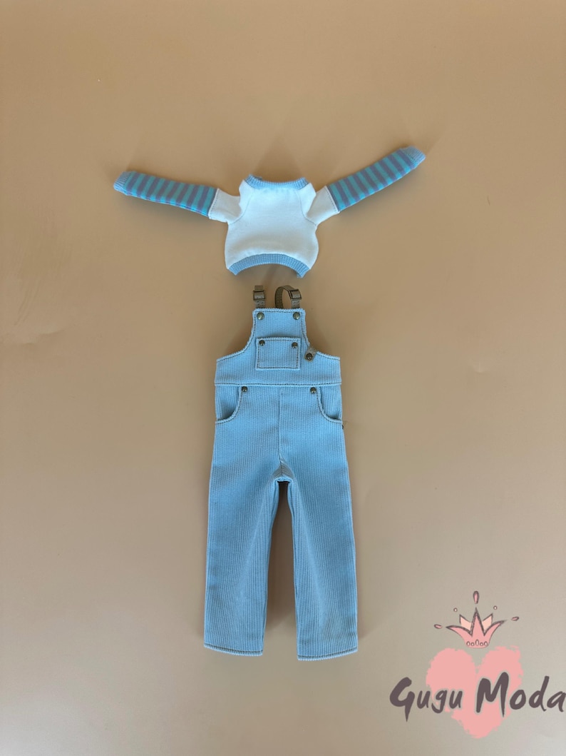 May include: A light blue corduroy overall set with a white and blue striped long-sleeve shirt. The overall has pockets and adjustable straps. The set is displayed on a tan background. The text "Gugu Moda" is in the bottom right corner.