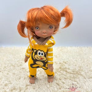 May include: A doll with orange hair in pigtails wearing a yellow and white striped jumpsuit with a monkey graphic. The doll has blue eyes and a light brown complexion.