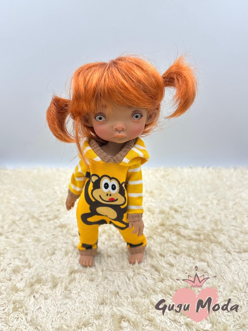May include: A doll with orange hair in pigtails wearing a yellow and white striped jumpsuit with a monkey graphic. The doll has blue eyes and a light brown complexion.