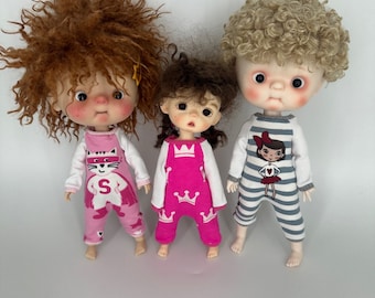 May include: Three dolls wearing pink and blue pajamas with different graphic designs. The dolls have curly hair and are standing on a white background.