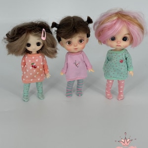 May include: Three small dolls wearing pajamas. The doll on the left has brown hair and a pink cherry-printed top and mint green leggings. The middle doll has brown pigtails and a pink top with a butterfly design and striped leggings. The doll on the right has pink and blonde hair and a mint green top with heart prints and pink leggings.
