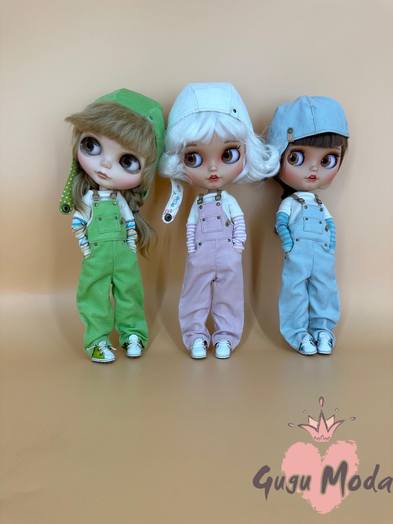 May include: Three dolls wearing overalls and hats. The dolls are dressed in green, pink, and blue overalls, with matching hats. Each doll wears a striped shirt and white sneakers. The dolls have large eyes and are posed in a row.