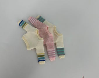 Puede incluir: Tres suéteres de muñeca en miniatura en colores crema, rosa y crema. Los suéteres crema tienen mangas a rayas en azul, verde y amarillo. El suéter rosa tiene mangas a rayas rosas y blancas. El logotipo de Gugu Moda está en la esquina inferior derecha.