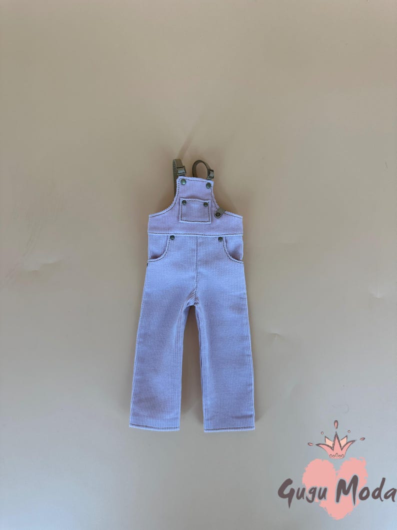 May include: Light purple corduroy overalls with adjustable straps and pockets. The overalls have a classic design with metal hardware. The Gugu Moda logo is in the bottom right corner.