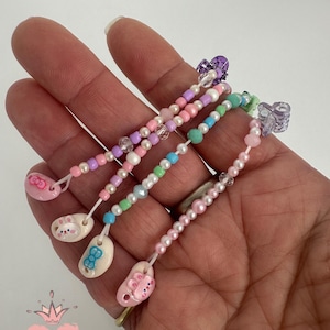May include: Four beaded pacifier clips in pastel colors, held in a hand. Each clip features a different design: a pink shoe with a bunny, a white shoe with a blue bow, a pink shoe with a cat, and a pink shoe with a cat. The clips are adorned with beads and clear plastic clasps.
