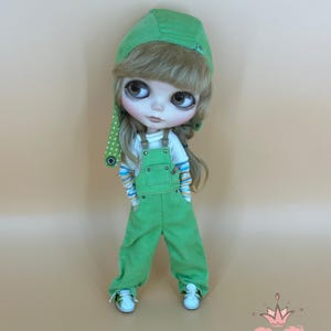 May include: A doll wearing a green corduroy overall outfit, a white shirt with colorful striped sleeves, and white sneakers. The doll has a green cap with a polka dot accent. The doll has long blonde hair and large eyes. The doll is standing in front of a light brown background.