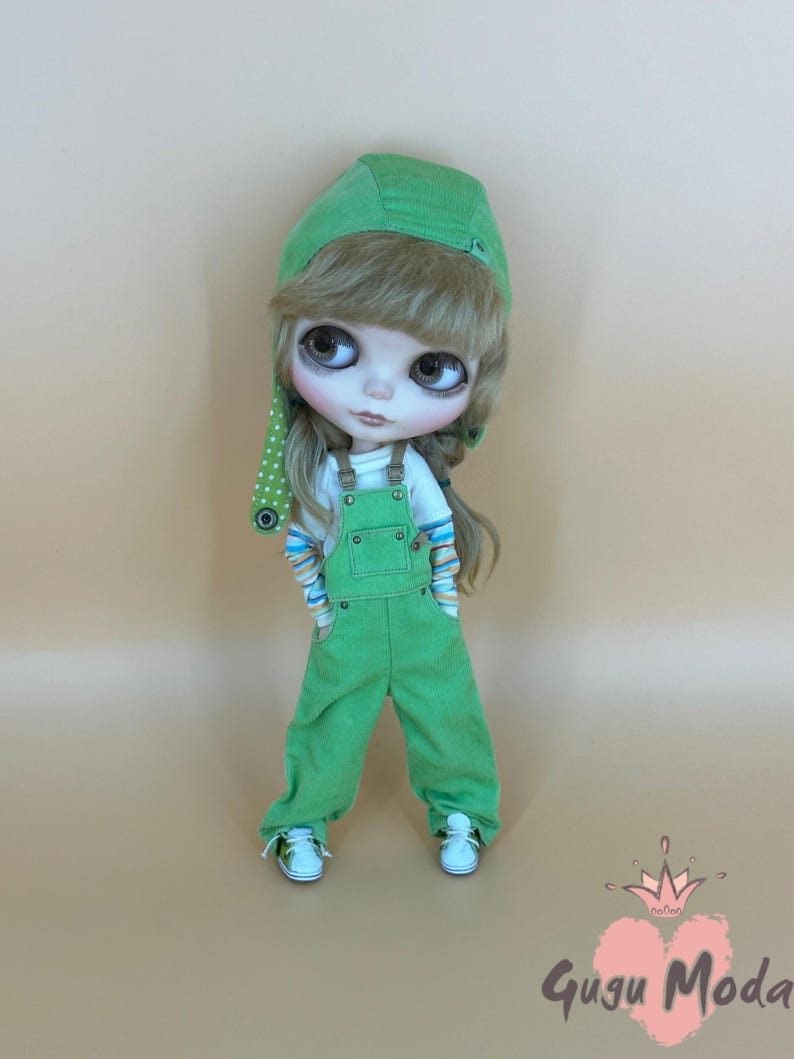 May include: A doll wearing a green corduroy overall outfit, a white shirt with colorful striped sleeves, and white sneakers. The doll has a green cap with a polka dot accent. The doll has long blonde hair and large eyes. The doll is standing in front of a light brown background.
