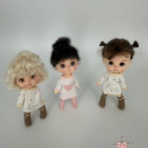 May include: Three small dolls wearing white dresses with brown boots. The dolls have different hair colors: blonde, black, and brown. The doll in the middle is wearing a pink heart on her dress.