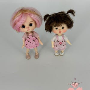 May include: Two small dolls with large eyes and colorful hair. One doll has pink and blonde hair, the other has brown hair in pigtails. Both dolls wear pink overalls with white shirts and matching shoes. The dolls are standing on a light gray surface.