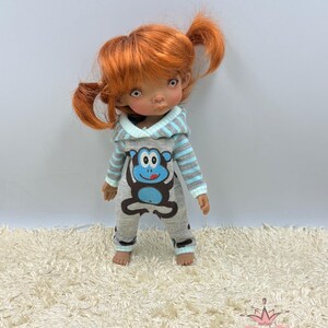 May include: A doll wearing a gray and blue hooded jumpsuit with a monkey graphic. The doll has red hair in pigtails and brown eyes.