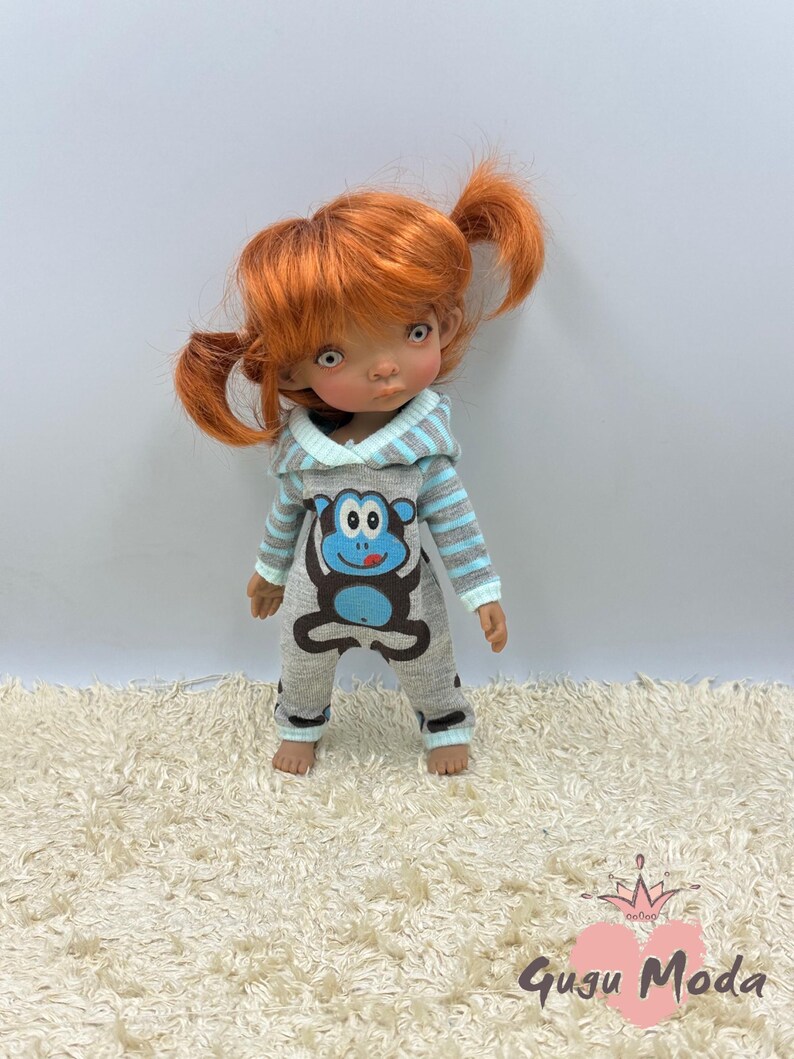 May include: A doll wearing a gray and blue hooded jumpsuit with a monkey graphic. The doll has red hair in pigtails and brown eyes.