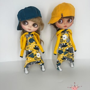May include: Two dolls wearing yellow and blue outfits with a fish and floral print. The dolls are wearing yellow and blue baseball caps. The dolls are standing on a white background.