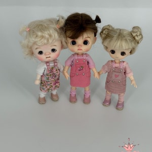 May include: Three small dolls with different hairstyles and outfits. The doll on the left has blonde curly hair and a floral print dress. The middle doll has brown hair and a pink dress with a cherry design. The doll on the right has blonde hair in buns and a pink dress with a heart design. The dolls are standing together.