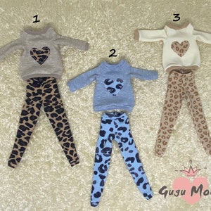 Puede incluir: Tres conjuntos de ropa para muñecas, cada uno con una camisa de manga larga y leggings. Las camisas son de color beige, azul claro y blanco con un diseño de corazón de estampado de leopardo. Los leggings son todos de estampado de leopardo.