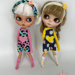 May include: Two dolls wearing colorful outfits and headbands. The doll on the left is wearing a pink and blue leopard print jumpsuit with a pink heart and pink socks. The doll on the right is wearing a blue and white daisy print jumpsuit with a yellow heart and yellow socks.