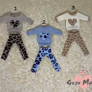 May include: Three sets of doll clothes featuring a long-sleeved sweater with a heart-shaped design and leggings. The sweaters are in beige, light blue, and white. The leggings are in a leopard print pattern with brown, black, and white colors.