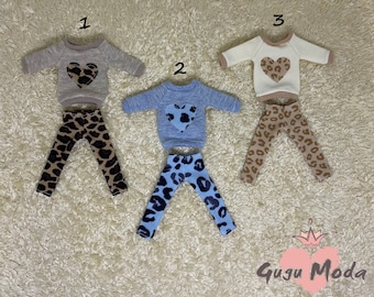 May include: Three sets of doll clothes featuring a long-sleeved sweater with a heart-shaped design and leggings. The sweaters are in beige, light blue, and white. The leggings are in a leopard print pattern with brown, black, and white colors.