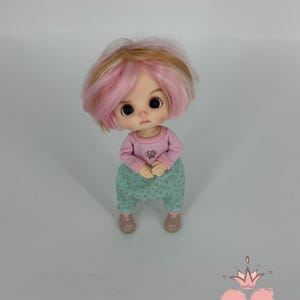 May include: A doll with pink and blonde hair, wearing a pink long-sleeved shirt and mint green pants with a floral pattern. The doll is wearing pink shoes. The doll has a sad expression. The Gugu Moda logo is in the bottom right corner.