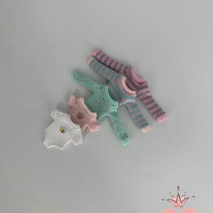 May include: A collection of doll clothes, including white, pink, and mint green tops and pajamas. The white top features a yellow bee graphic. The pajamas have striped and dotted patterns. The Gugu Moda logo is in the bottom right corner.