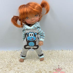 May include: A doll wearing a grey and blue hooded jumpsuit with a monkey graphic. The doll has red hair in pigtails and brown eyes.