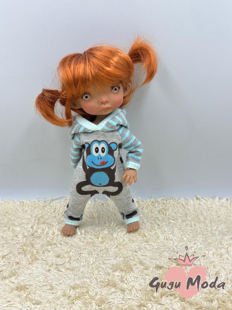 May include: A doll wearing a grey and blue hooded jumpsuit with a monkey graphic. The doll has red hair in pigtails and brown eyes.