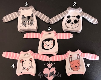 May include: Five different doll sweaters with pink and grey stripes. Each sweater has a different animal graphic on the front: a cat, a panda, a sheep, a mouse, and a rabbit.