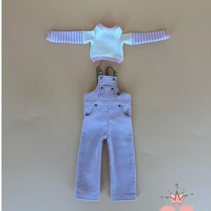 May include: A doll's outfit featuring a white long-sleeved shirt with pink striped sleeves and a pink corduroy overall. The overall has adjustable straps and pockets. The outfit is displayed on a neutral background. The text "Gugu Moda" is visible.