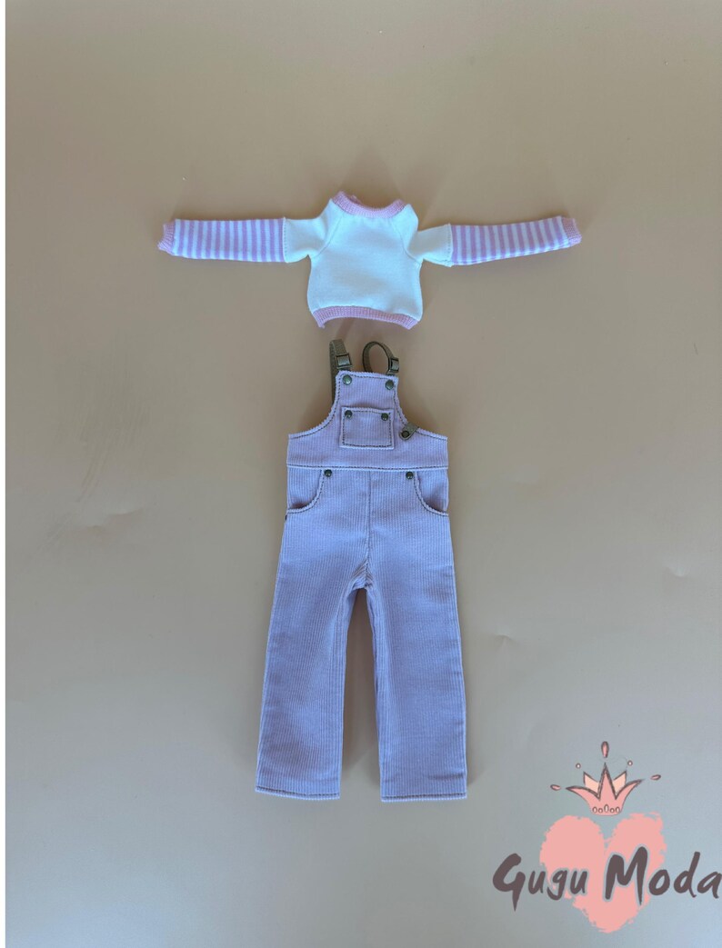 May include: A doll's outfit featuring a white long-sleeved shirt with pink striped sleeves and a pink corduroy overall. The overall has adjustable straps and pockets. The outfit is displayed on a neutral background. The text "Gugu Moda" is visible.