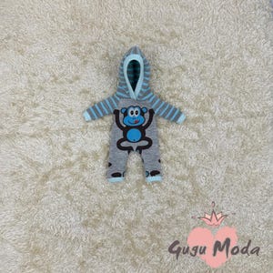 May include: A gray and blue hooded baby onesie with a monkey design. The onesie has a blue monkey with a blue tongue and white eyes. The onesie has a hood with blue and white stripes.