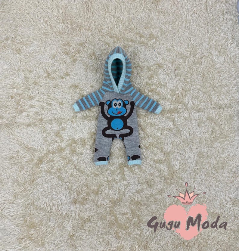 May include: A gray and blue hooded baby onesie with a monkey design. The onesie has a blue monkey with a blue tongue and white eyes. The onesie has a hood with blue and white stripes.