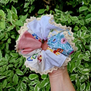 Könnte beinhalten: Ein Set aus fünf Stoff-Haargummis mit gestickten Blumenmustern. Die Haargummis sind aus weißem, rosa und blauem Stoff und haben einen weißen Spitzenbesatz.