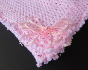 May include: A soft pink crocheted baby blanket with a delicate openwork pattern and scalloped edges. A light pink satin ribbon bow is tied at the corner, adding a touch of elegance. The blanket is perfect for a baby shower gift.