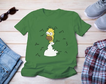 May include: A green t-shirt featuring a cartoon image of a yellow character with a beard, peeking out from behind a bush. The character is wearing a white shirt and has a surprised expression.