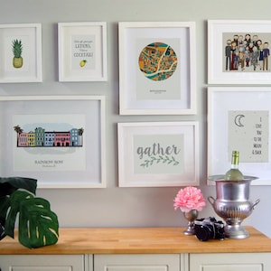 May include: A gallery wall with white frames featuring various prints, including a pineapple, a lemon, a city map, a family portrait, a moon and stars design, a floral design, and a cityscape. The wall is painted a light gray color. A wooden countertop with a silver ice bucket and a bottle of wine is in the foreground.