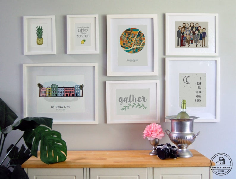 May include: A gallery wall with white frames featuring various prints, including a pineapple, a lemon, a city map, a family portrait, a moon and stars design, a floral design, and a cityscape. The wall is painted a light gray color. A wooden countertop with a silver ice bucket and a bottle of wine is in the foreground.
