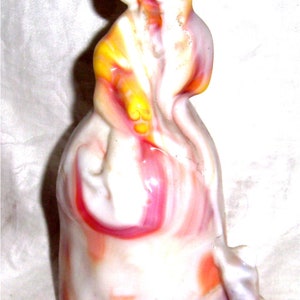 May include: A vintage glass figurine of a woman in a white, pink, and yellow swirl pattern. The figurine is in a standing pose with her arms crossed.