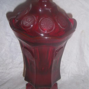 May include: A red glass candy dish with a lid. The dish has a scalloped edge and is decorated with raised circles. The lid has a knob on top.